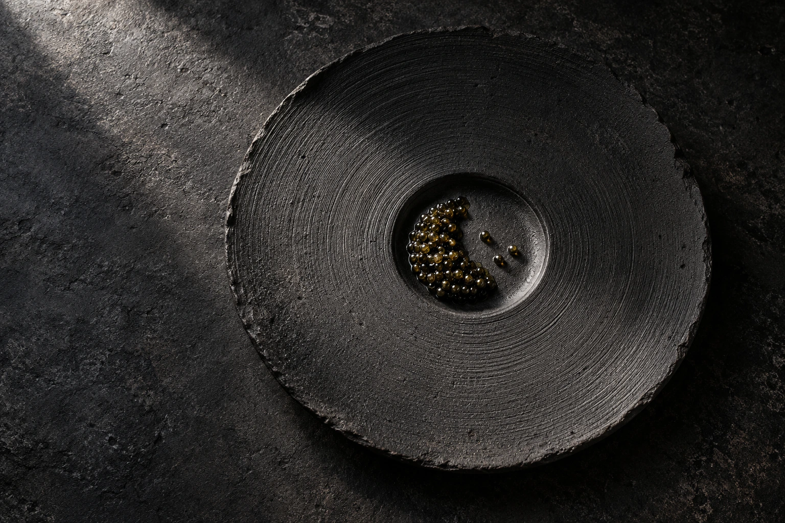 The complete still-life photograph of caviar on a sculptural black ceramic plate with delicate concentric grooves